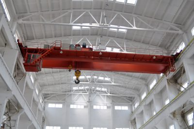 Top-running red overhead bridge crane with hoist inside a bright industrial bay, illustrating overhead crane maintenance and inspection.
